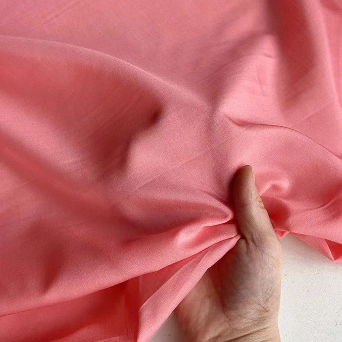 Une main rassemble délicatement un morceau de Soie de coton multicolore au mètre de Longan Craft France, créant des plis doux et des ombres sur la doublure en soie respirante. Le fond est uni et de couleur claire.