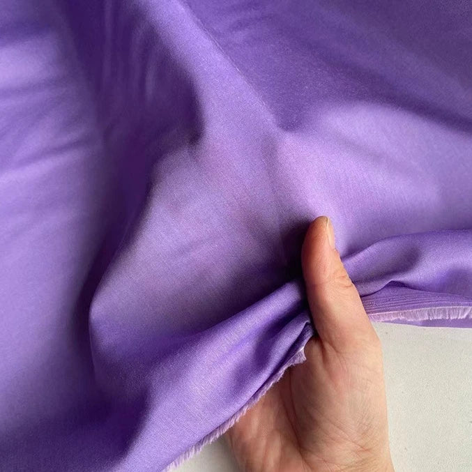 Une main tient un morceau de Soie de coton multicolore au mètre de Longan Craft France - ce tissu de soie de mûrier doublure, léger et respirant, présente un léger éclat et une texture douce lorsque le reste est posé à plat sur la surface.