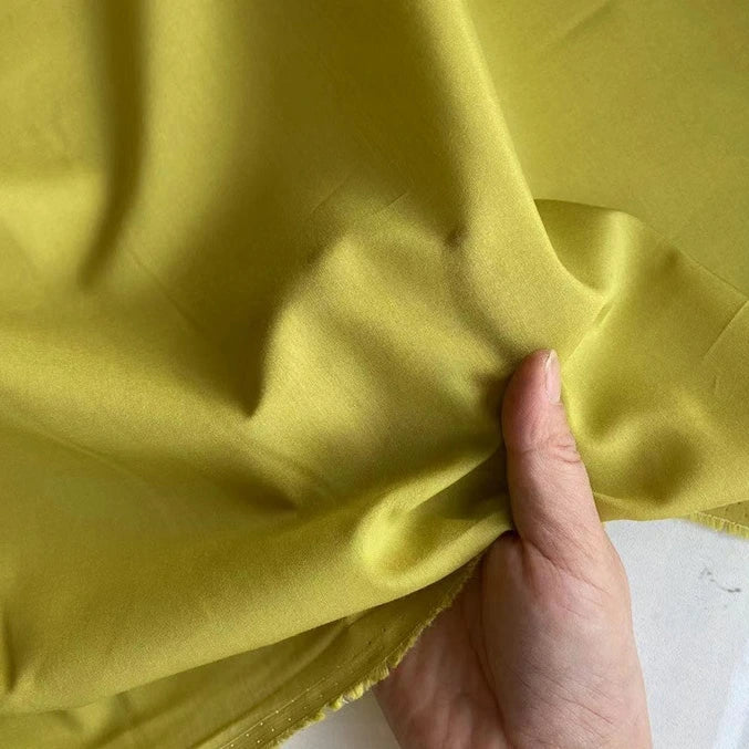 Une main tient le Soie de coton multicolore au mètre de Longan Craft France, un tissu de doublure en soie de mûrier léger et respirant, de couleur jaune-vert clair, montrant sa texture lisse et sa brillance subtile avec des plis naturels.