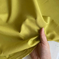 Une main tient le Soie de coton multicolore au mètre de Longan Craft France, un tissu de doublure en soie de mûrier léger et respirant, de couleur jaune-vert clair, montrant sa texture lisse et sa brillance subtile avec des plis naturels.