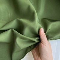 Une main tient un morceau de Soie de coton multicolore au mètre de Longan Craft France - un tissu de soie mauve, lisse, doux, mat et respirant - légèrement froncé et soulevé sur un arrière-plan flou.