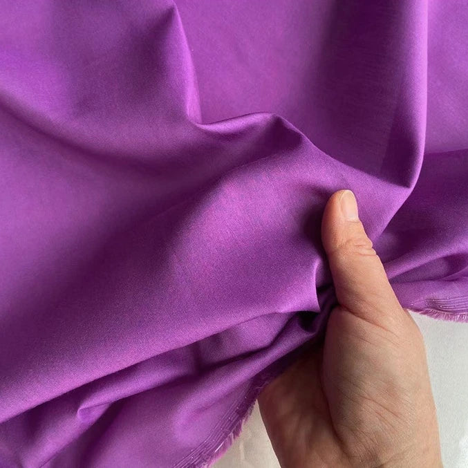 Une main soulève un morceau de Soie de coton multicolore au mètre, lisse et brillant, de Longan Craft France, mettant en valeur la texture et le drapé délicats de ce tissu de doublure en soie de mûrier, léger et respirant.