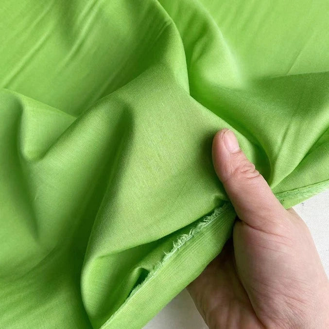 Une main tient un morceau de Soie de coton multicolore au mètre de Longan Craft France, mettant en valeur sa texture lisse, sa couleur vert vif et son bord non fini - un tissu de doublure en soie léger et respirant.