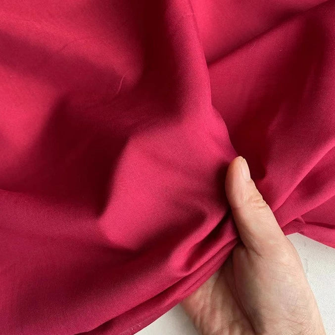 Une main rassemble un morceau de Soie de coton multicolore rose vif au mètre de Longan Craft France, mettant en valeur sa texture douce et légère et sa brillance subtile sur un fond de tissu assorti avec une touche de blanc.