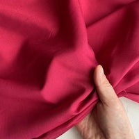 Une main rassemble un morceau de Soie de coton multicolore rose vif au mètre de Longan Craft France, mettant en valeur sa texture douce et légère et sa brillance subtile sur un fond de tissu assorti avec une touche de blanc.