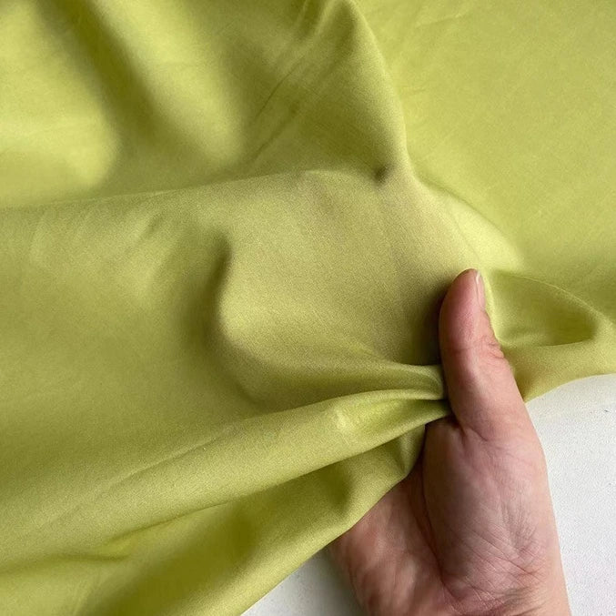 Une main pince un morceau de Soie de coton multicolore au mètre de Longan Craft France, montrant la texture lisse et légèrement brillante de ce tissu de doublure en soie de mûrier, léger et respirant, étalé sur une surface plane.
