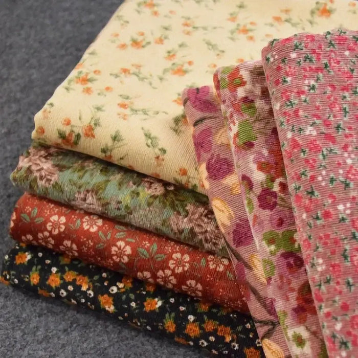 Une pile de six Tissu velours côtelé à fleurs colorées au mètre de Longan Craft France, chacun avec des motifs floraux uniques et vibrants, est disposée sur une surface texturée grise - parfaite pour la confection, l'ameublement et les loisirs créatifs.