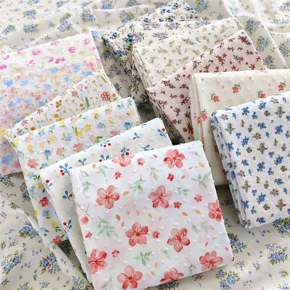 Des carrés pliés de Tissu plumetis blanc imprimé de fleurs de Longan Craft France, un tissu de plumetis de coton à motifs floraux, sont présentés sur une surface de tissu floral assorti.