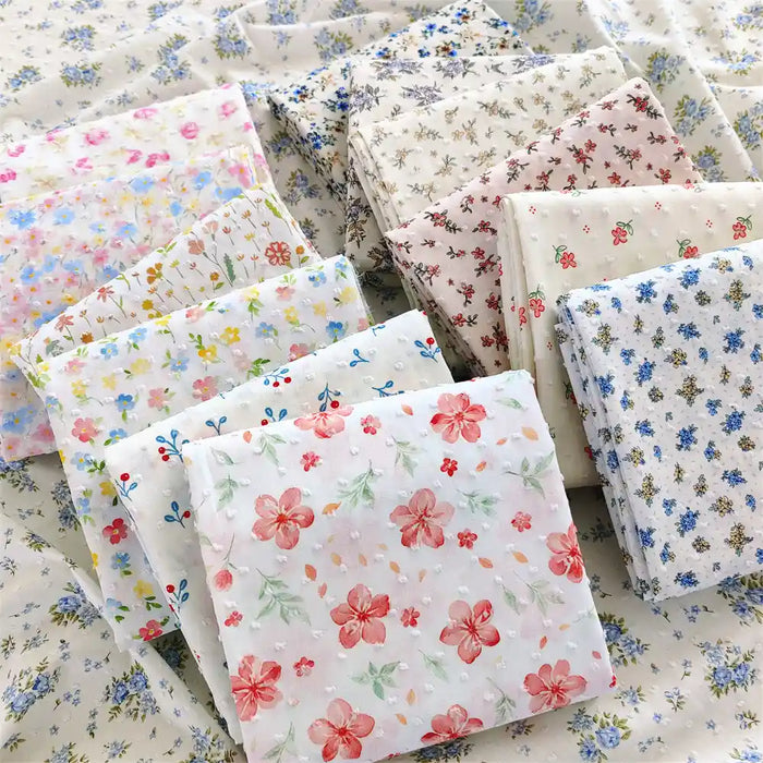 Des carrés pliés de Tissu plumetis blanc imprimé de fleurs de Longan Craft France, un tissu de plumetis de coton à motifs floraux, sont présentés sur une surface de tissu floral assorti.