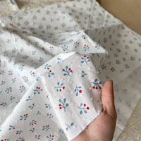 Une main tient un morceau de Tissu plumetis blanc imprimé de fleurs au mètre de Longan Craft France ; en arrière-plan, plus de ce tissu en coton plumetis à motif floral est étalé sur une surface plane.