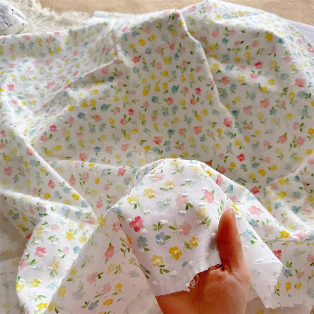 Une main tient un morceau de Tissu plumetis blanc imprimé de fleurs par Longan Craft France, un tissu de coton fin et semi-transparent avec des motifs floraux colorés, drapé sur une surface plane - idéal pour les vêtements d'intérieur.