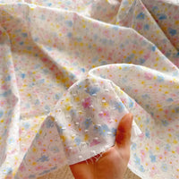 Une main tient un morceau de tissu plumetis blanc imprimé de fleurs au mètre par Longan Craft France, avec son motif floral pastel en rose, bleu, jaune et violet. Le tissu de coton léger est étalé derrière pour montrer sa texture délicate.