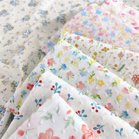 Plusieurs pièces de Tissu plumetis blanc imprimé de fleurs de Longan Craft France sont superposées et présentent des motifs floraux et botaniques pastel, certains mettant en valeur des fleurs bleues, roses et jaunes tandis que d'autres présentent des points texturés ou des motifs feuillus.