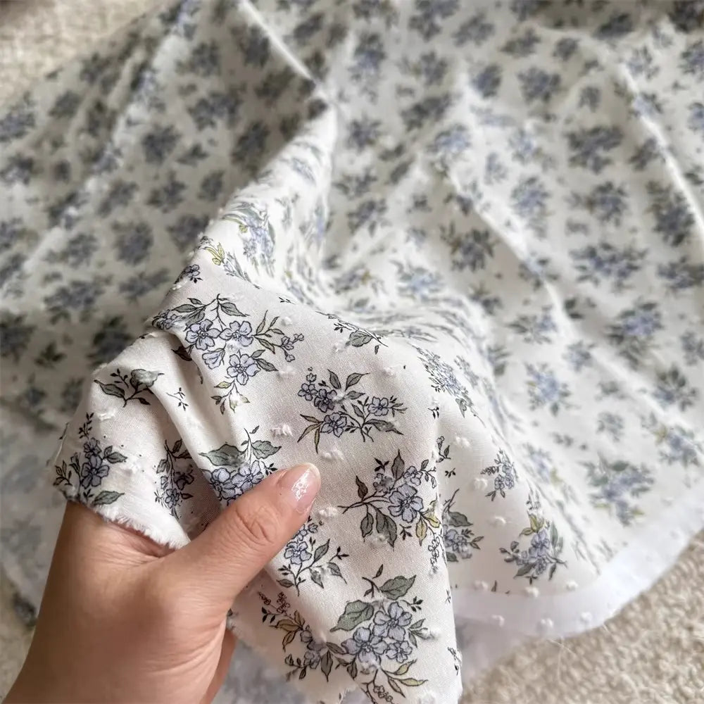 Une main tient un morceau de Tissu plumetis blanc imprimé de fleurs au mètre par Longan Craft France, un tissu de coton avec un motif floral, tandis que d'autres pièces du même textile sont étalées à l'arrière-plan.