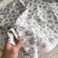 Une main tient un morceau de Tissu plumetis blanc imprimé de fleurs au mètre par Longan Craft France, un tissu de coton avec un motif floral, tandis que d'autres pièces du même textile sont étalées à l'arrière-plan.