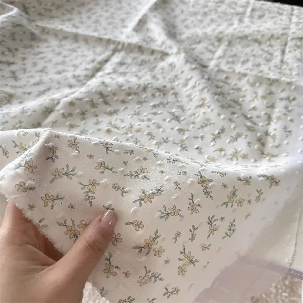Une main présente le Tissu plumetis blanc imprimé de fleurs de Longan Craft France, un tissu de coton aux délicats motifs floraux, tandis que d'autres pièces de cet élégant tissu en coton plumetis sont disposées à l'arrière-plan.