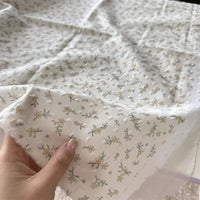 Une main présente le Tissu plumetis blanc imprimé de fleurs de Longan Craft France, un tissu de coton aux délicats motifs floraux, tandis que d'autres pièces de cet élégant tissu en coton plumetis sont disposées à l'arrière-plan.