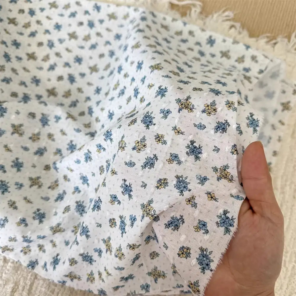 Une main tient Tissu plumetis blanc imprimé de fleurs par Longan Craft France, un tissu de coton avec un délicat motif floral et des bords effilochés, montré légèrement froissé sur une surface tissée légère.