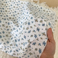 Une main tient Tissu plumetis blanc imprimé de fleurs par Longan Craft France, un tissu de coton avec un délicat motif floral et des bords effilochés, montré légèrement froissé sur une surface tissée légère.