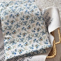 Un morceau de Tissu plumetis blanc imprimé de fleurs par Longan Craft France repose plié sur un tissu beige avec texte noir, tous deux placés à l'intérieur d'un panier rectangulaire tressé.