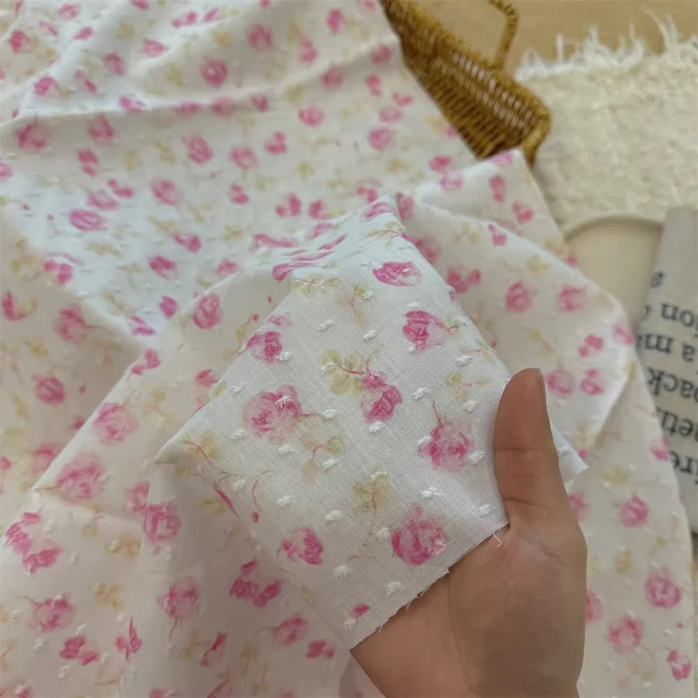 Une main tient le tissu plumetis blanc imprimé de fleurs au mètre de Longan Craft France, un tissu de coton avec des fleurs roses et des points texturés, assorti à la pièce plus grande ci-dessous, avec un panier tressé et une natte à franges à l'arrière-plan.