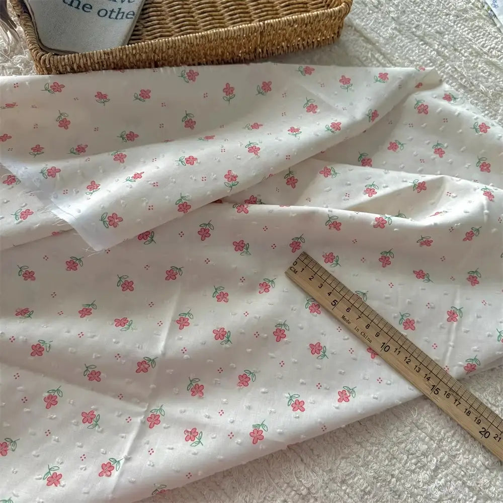 Une longueur de tissu plumetis blanc imprimé de fleurs (coton) de Longan Craft France, présentant un délicat motif floral, est exposée sur une surface texturée, avec une règle en bois et un panier tressé à proximité.