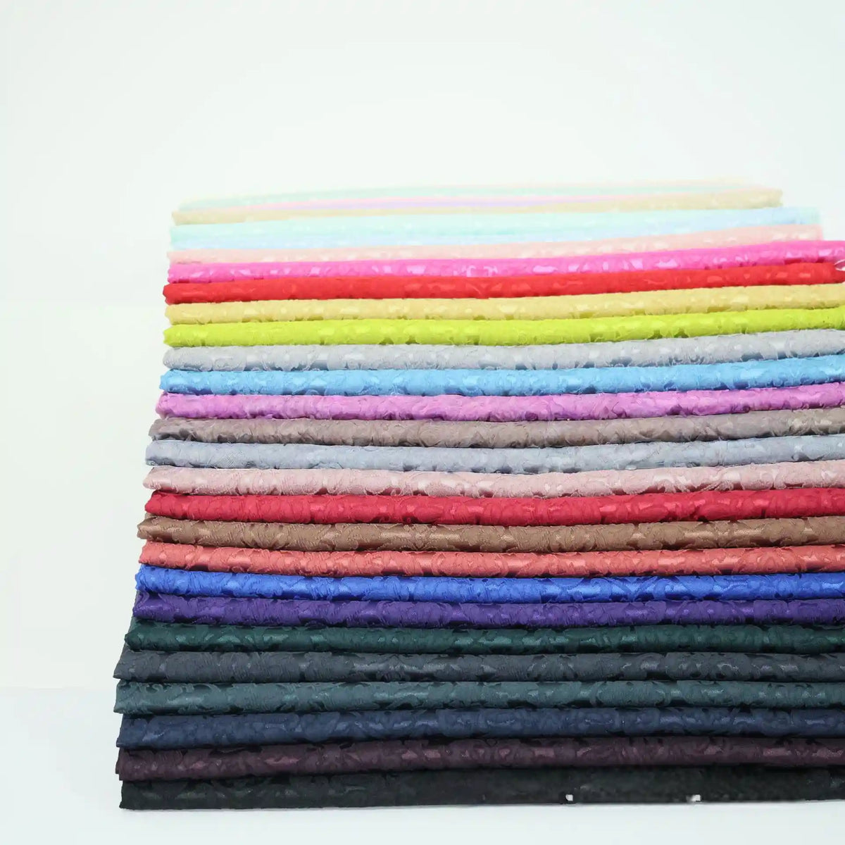 Une pile de tissu damassé jacquard multicolore de Longan Craft France, avec des motifs floraux, est soigneusement pliée en couches sur un fond blanc.