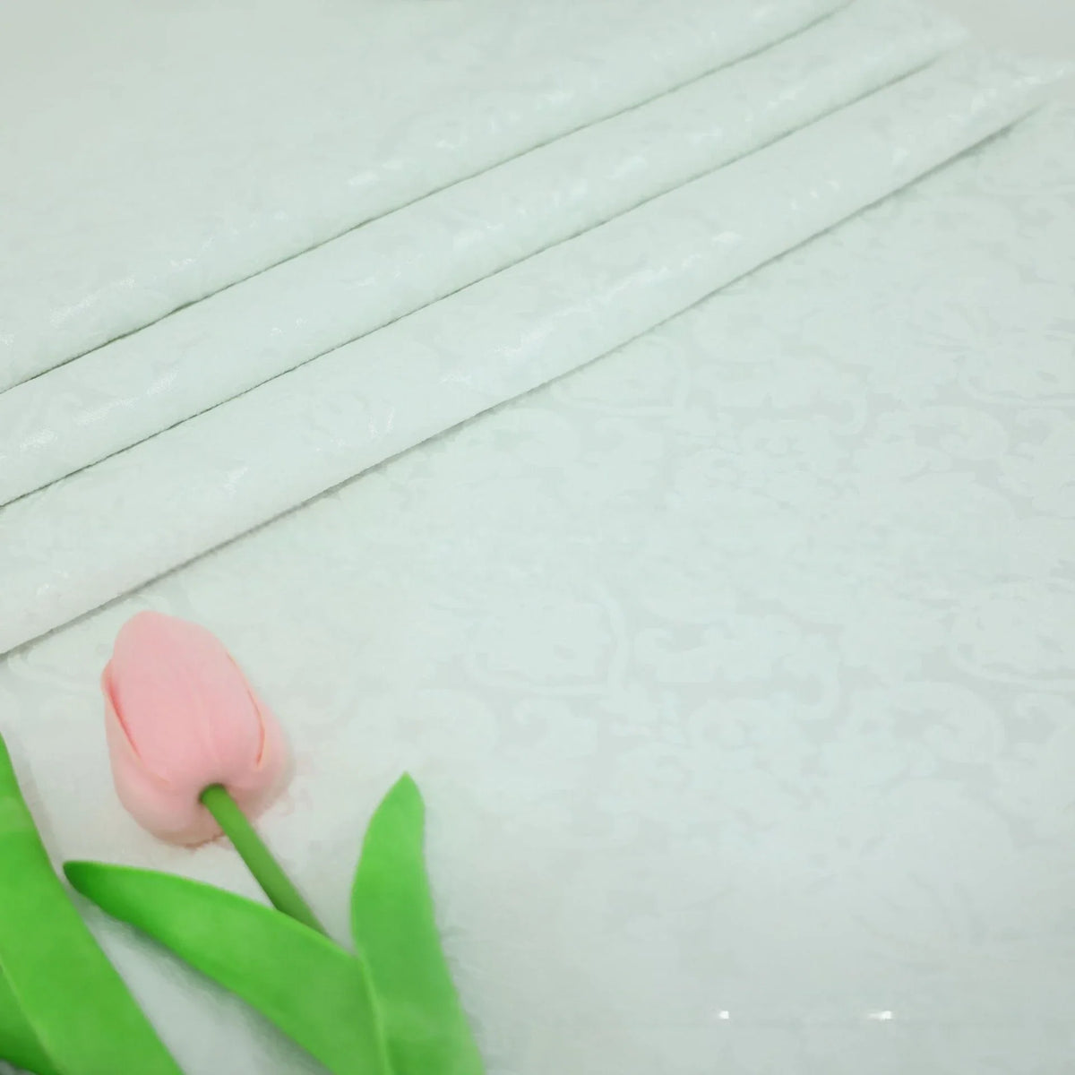Une tulipe rose repose sur un Tissu jacquard damassé multicolore à motif floral de Longan Craft France, avec trois pièces pliées du même tissu exposées dans un coin.