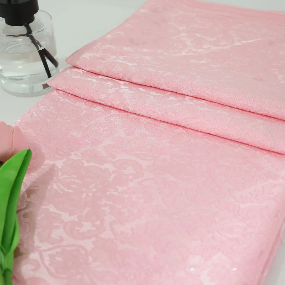 Un morceau plié de tissu damassé jacquard à motif floral de Longan Craft France repose sur une surface blanche à côté d'une fleur artificielle verte et d'un flacon pulvérisateur transparent.