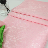 Un morceau plié de tissu damassé jacquard à motif floral de Longan Craft France repose sur une surface blanche à côté d'une fleur artificielle verte et d'un flacon pulvérisateur transparent.