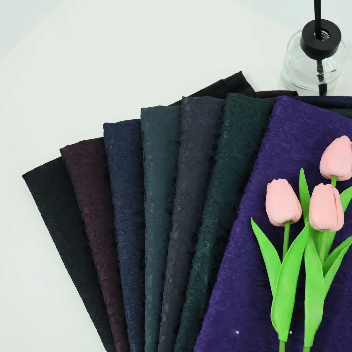 Huit tissus d'ameublement en damas jacquard de Longan Craft France sont disposés en éventail sur une surface blanche. À proximité, trois tulipes artificielles roses et un vase à couvercle noir complètent le textile floral multicolore.
