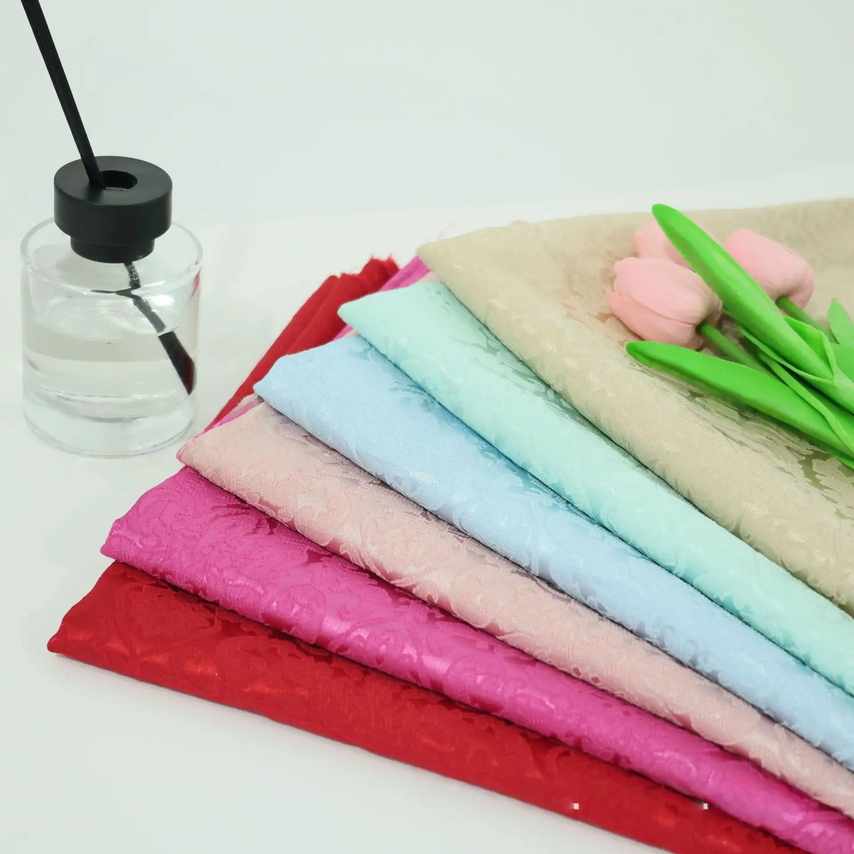 Plusieurs pièces pliées de tissu d'ameublement jacquard damassé de Longan Craft France aux motifs floraux multicolores sont empilées à côté d'un pot transparent à couvercle noir et de deux tulipes roses artificielles, le tout présenté sur une surface blanche.