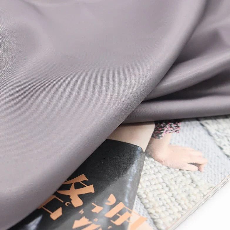 Un gros plan du Tissu satiné coloré en viscose au mètre de Longan Craft France, un tissu polyester-viscose sergé gris clair et lisse, couvre partiellement une page de magazine avec un texte japonais et une photo ; la texture de la moquette apparaît à l'arrière-plan.