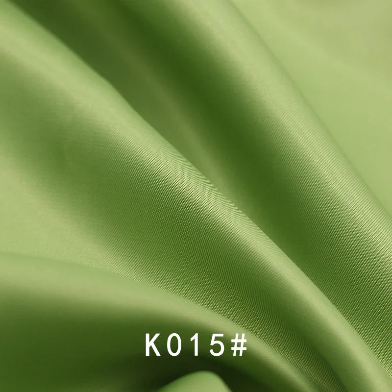 Gros plan sur le Tissu satiné coloré en viscose au mètre de Longan Craft France, montrant sa texture vert clair, lisse et légèrement brillante avec de légers plis. Le code "K015#" est indiqué en texte blanc au bas de l'image.