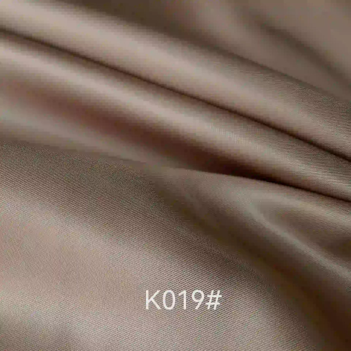 Gros plan sur le Tissu satiné coloré en viscose au mètre de Longan Craft France, lisse et marron clair, avec de légers plis. Le code "K019#" apparaît en blanc en bas au centre, mettant en valeur cette élégante étoffe idéale pour robes de cérémonie.