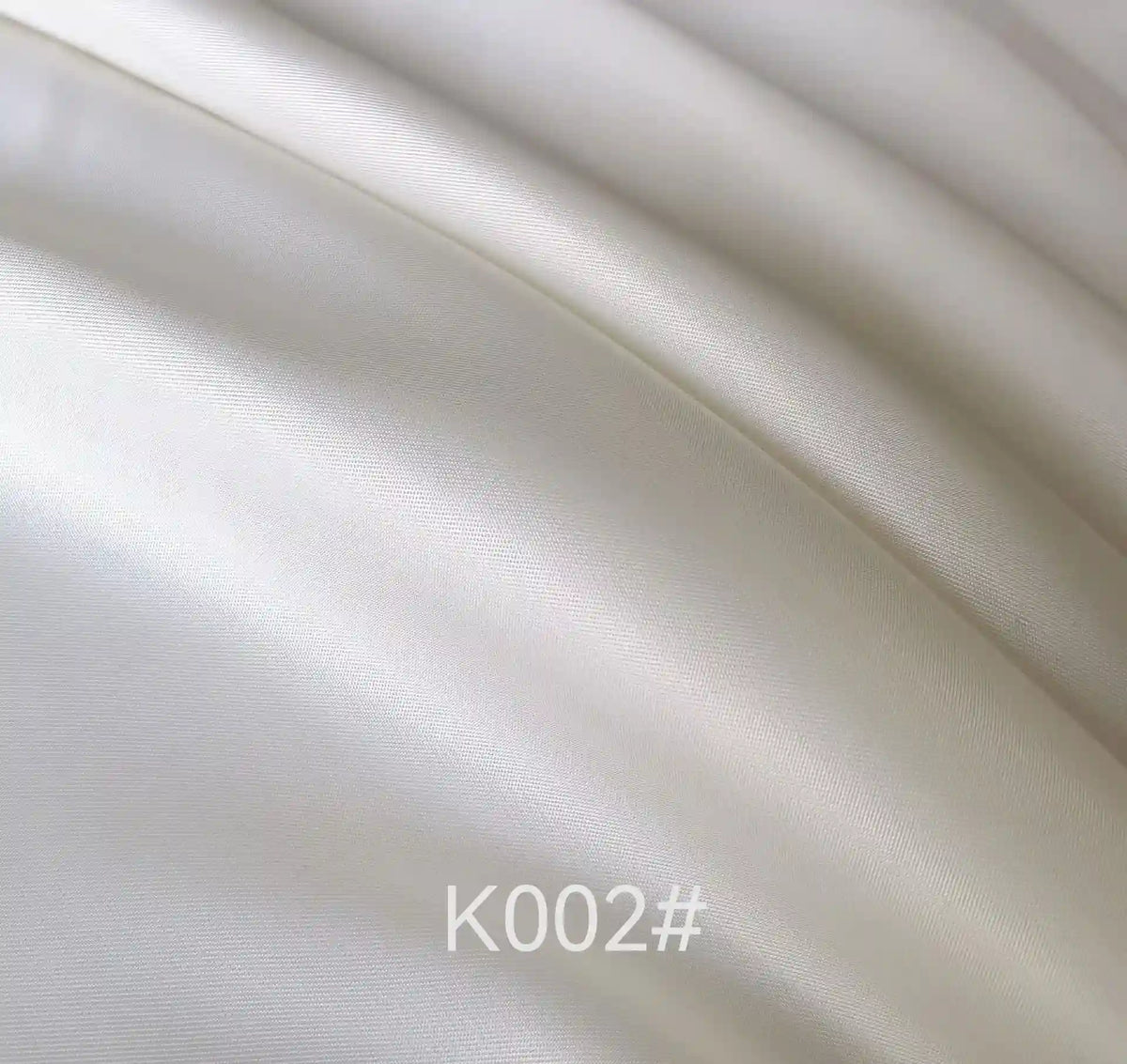 Gros plan sur le "Tissu satiné coloré en viscose au mètre" lisse et brillant de Longan Craft France, montrant sa brillance élégante et ses plis délicats. Le code "K002#" apparaît en bas au centre, mettant en valeur ce tissu en viscose-polyester pour robes.