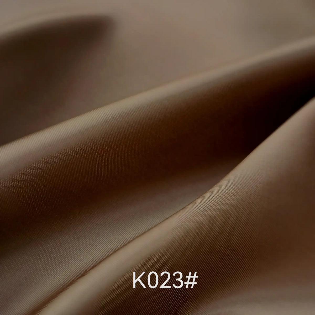 Gros plan sur un tissu satiné en viscose brun et lisse de Longan Craft France, montrant de légers plis et une brillance subtile. La mention "K023#" apparaît en bas au centre. Idéal pour les robes de cérémonie ; vendu au mètre avec le tissage sergé polyester-viscose.