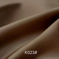 Gros plan sur un tissu satiné en viscose brun et lisse de Longan Craft France, montrant de légers plis et une brillance subtile. La mention "K023#" apparaît en bas au centre. Idéal pour les robes de cérémonie ; vendu au mètre avec le tissage sergé polyester-viscose.