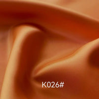 Gros plan sur un Tissu satiné coloré en viscose au mètre (Longan Craft France), lisse et orange, aux plis souples et à la brillance subtile. La mention "K026#" apparaît en blanc au centre de la partie inférieure, mettant en valeur cet élégant tissu pour robes de cérémonie.