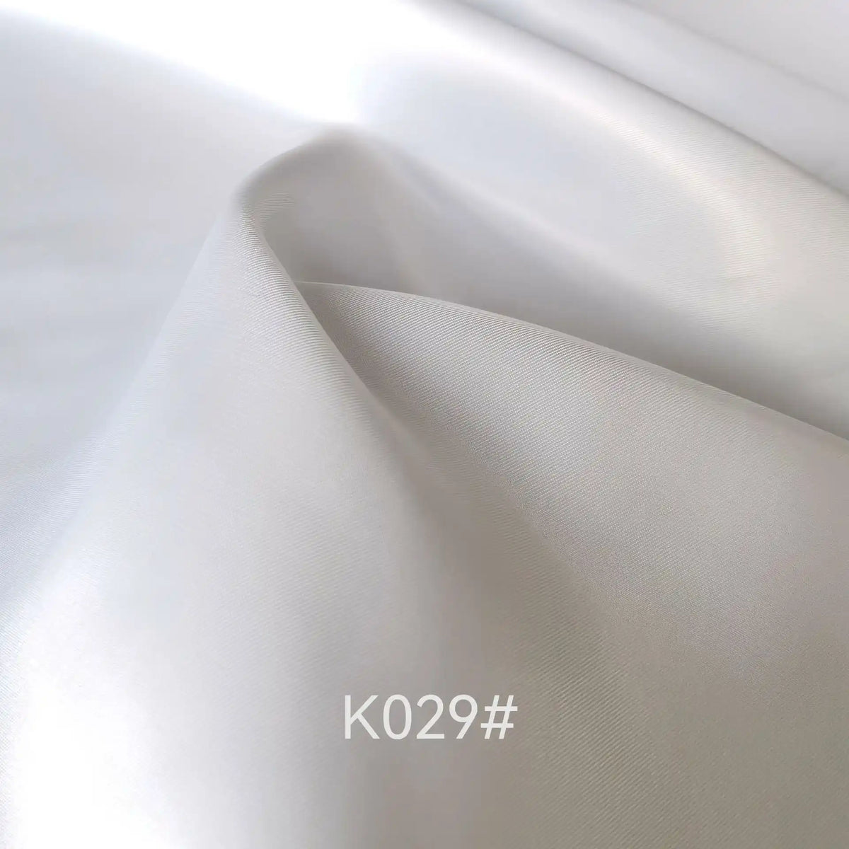 Gros plan sur un tissu blanc satiné en viscose de Longan Craft France, montrant de légers plis et un éclat doux. La mention "K029#" est imprimée en bas au centre. Idéal pour les robes de cérémonie, ce tissu polyester-viscose est vendu au mètre.