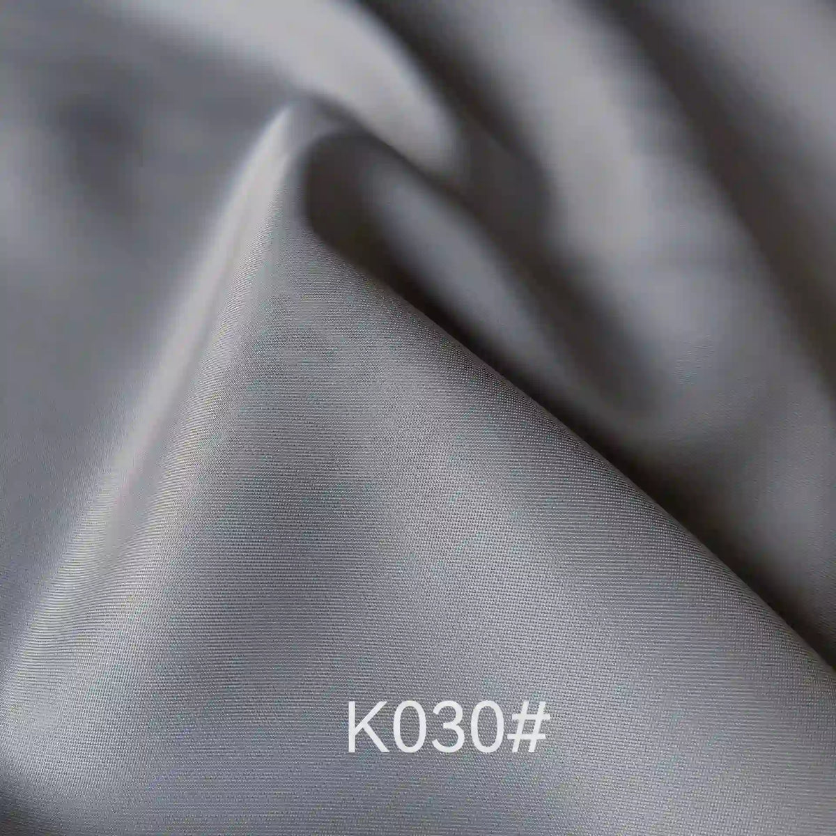 Gros plan sur le tissu satiné gris argenté lisse de Longan Craft France, montrant la texture élégante et les plis souples. La mention "K030#" figure en bas au centre. Idéal pour les robes de cérémonie.