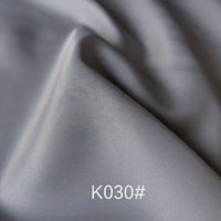 Gros plan sur le tissu satiné gris argenté lisse de Longan Craft France, montrant la texture élégante et les plis souples. La mention "K030#" figure en bas au centre. Idéal pour les robes de cérémonie.