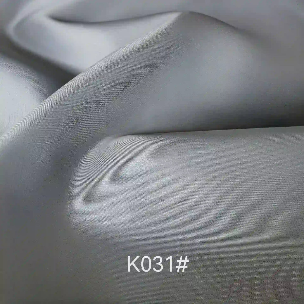 Gros plan sur un tissu de satin de viscose lisse et gris clair, dont la texture est visible et qui présente de légers plis. La mention "K031#" apparaît en blanc en bas au centre. Produit : Tissu satiné coloré en viscose au mètre par Longan Craft France, idéal pour les robes élégantes.