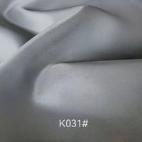 Gros plan sur un tissu de satin de viscose lisse et gris clair, dont la texture est visible et qui présente de légers plis. La mention "K031#" apparaît en blanc en bas au centre. Produit : Tissu satiné coloré en viscose au mètre par Longan Craft France, idéal pour les robes élégantes.