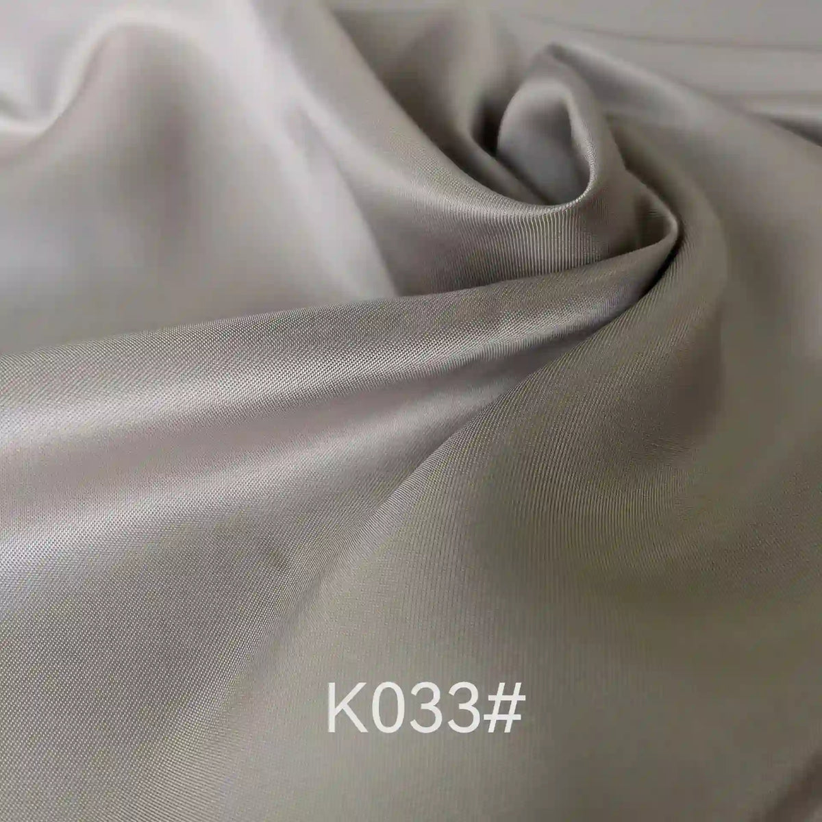 Gros plan sur un tissu satiné coloré en viscose au mètre, gris clair et lisse, de Longan Craft France, aux plis délicats et à l'éclat subtil. Le code "K033#" est superposé en texte blanc près du centre inférieur.