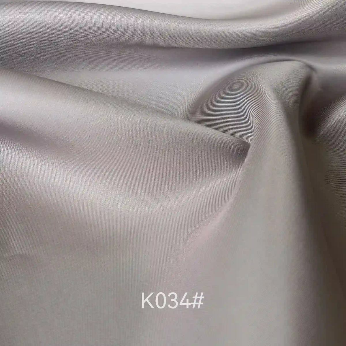 Gros plan sur le tissu satiné coloré en viscose au mètre de Longan Craft France, montrant sa surface lisse gris clair avec une brillance subtile et des plis. Le texte "K034#" apparaît en bas au centre. Idéal pour les robes élégantes et les cérémonies.