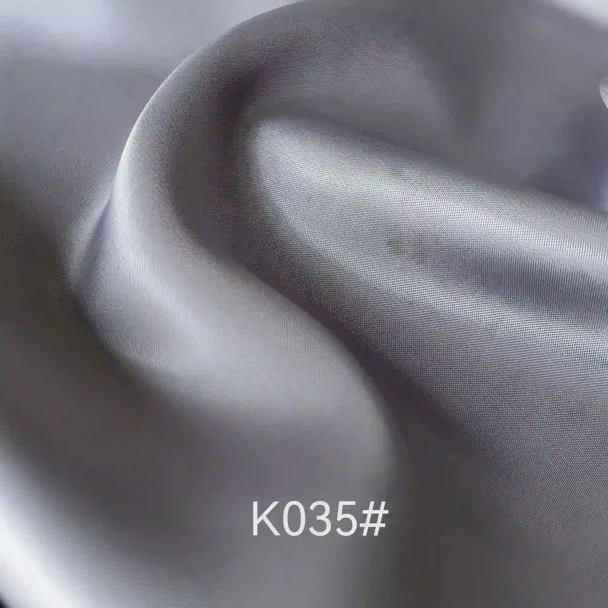 Gros plan sur le Tissu satiné coloré en viscose au mètre, gris clair et lisse, de Longan Craft France, montrant une texture fine et des plis souples. La mention "K035#" apparaît en bas au centre. Idéal pour les robes de cérémonie élégantes.