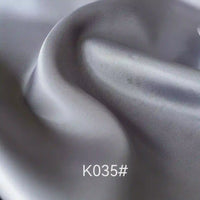 Gros plan sur le Tissu satiné coloré en viscose au mètre, gris clair et lisse, de Longan Craft France, montrant une texture fine et des plis souples. La mention "K035#" apparaît en bas au centre. Idéal pour les robes de cérémonie élégantes.