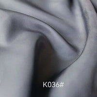 Gros plan sur un tissu satiné coloré en viscose au mètre, gris clair et lisse, de Longan Craft France, avec des plis et des ombres douces. La mention "K036#" apparaît en bas au centre en texte blanc. Idéal pour les robes de cérémonie élégantes, tissage sergé polyester-viscose.
