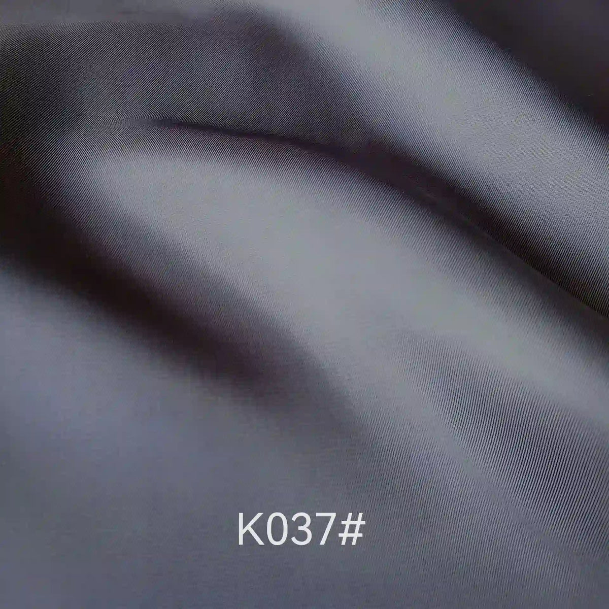 Élégant tissu satiné en viscose gris foncé plié de Longan Craft France, au tissage sergé et à la texture douce, parfait pour les robes de cérémonie. Le code "K037#" apparaît en blanc en bas au centre de l'image.